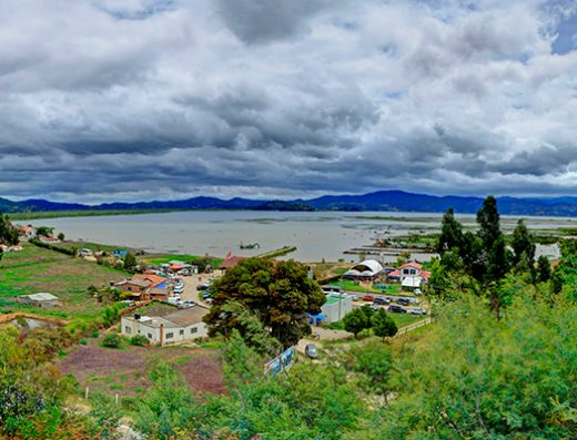 Vista de la Laguna de Fúquene con lanchas en el muelle, aves acuáticas sobre el agua y montañas verdes del altiplano cundiboyacense al fondo