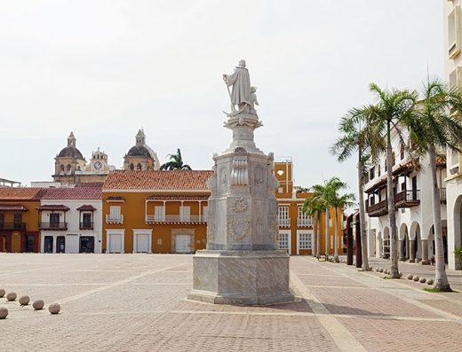 Plaza de la Aduana en Cartagena con estatua de Cristóbal Colón en el centro, Palacio de la Inquisición y Alcaldía al fondo, bajo cielo azul.